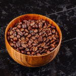 A close-up of medium roast coffee beans in a wooden bowl, elegantly placed on a black surface, showcasing their natural sheen.