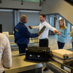 A man assists a woman by handing her a piece of luggage, showcasing teamwork in a commercial security setting.